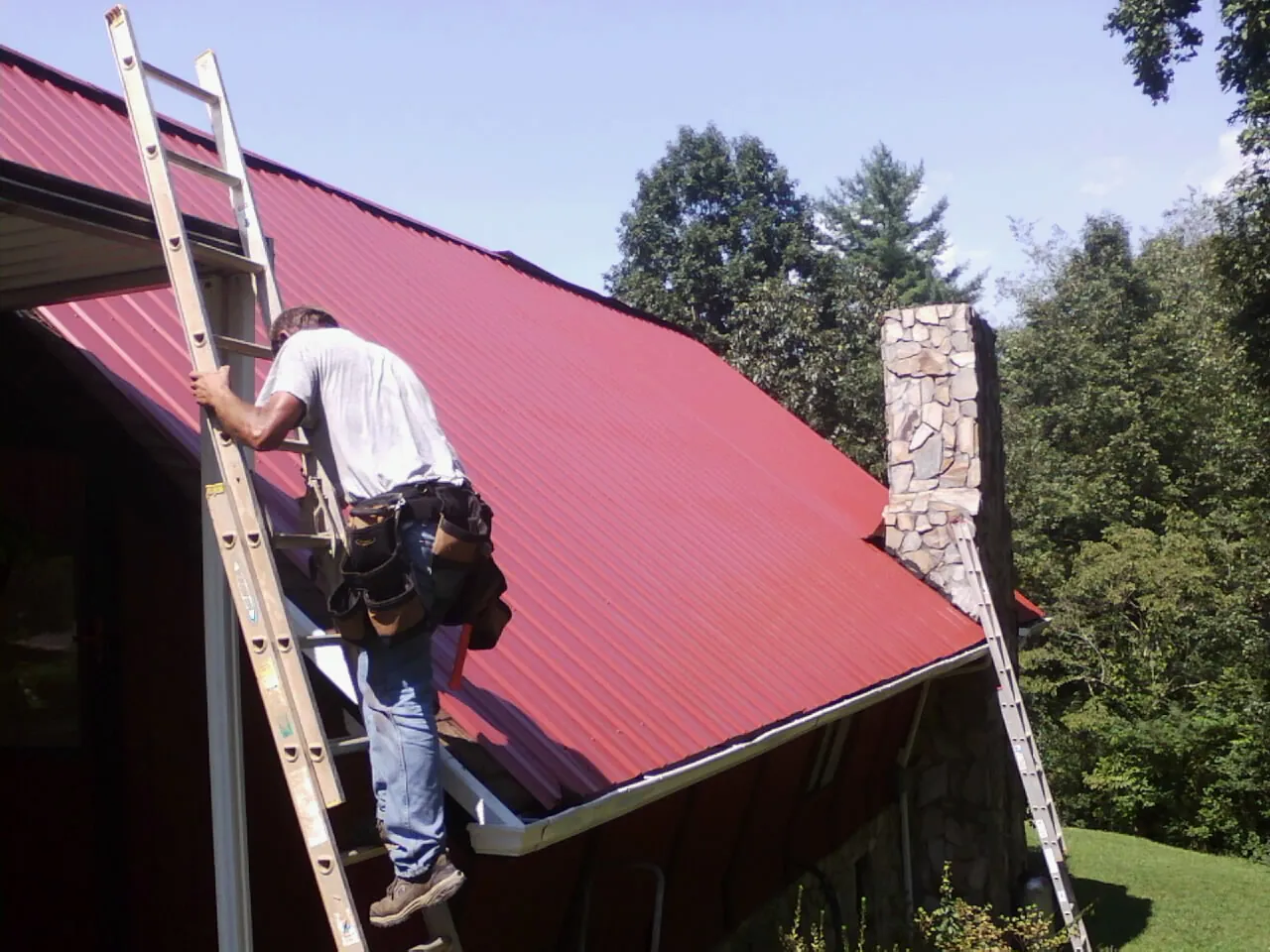 Roofer on a ladder beside a red metal roof and stone chimney — Soffit & Fascia Repair by Roofing Brothers in Palm Springs